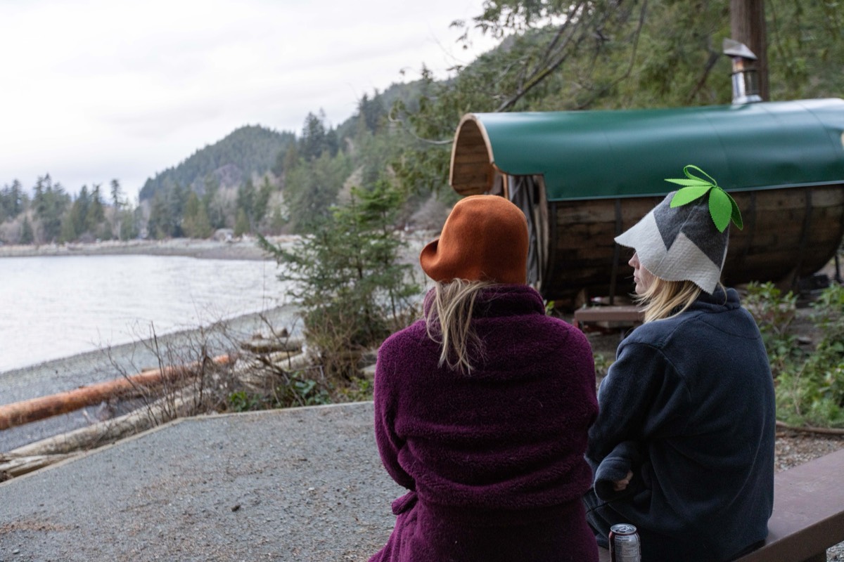 Two friends in felt sauna hats cooling down beside a wood-fired barrel sauna by the water